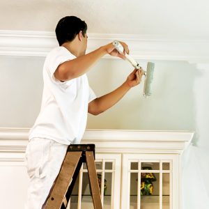 A house painter stands on a ladder to paint the upper portion of a room's wall.  RM